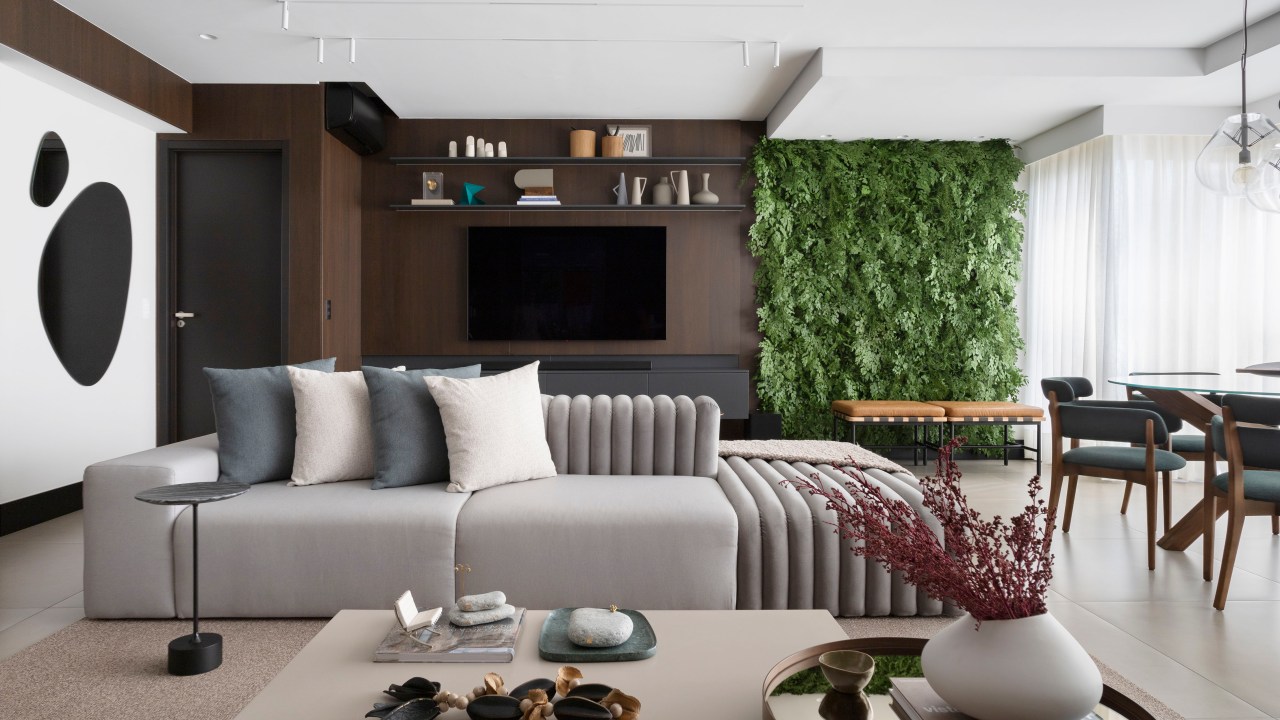 Sofá ilha é ponto central da sala de apê de 135m² com área gourmet. Projeto de Teto Arquitetura. Na foto, sala de estar com sofá ilha, jardim vertical e parede revestida com painel de madeira escura.