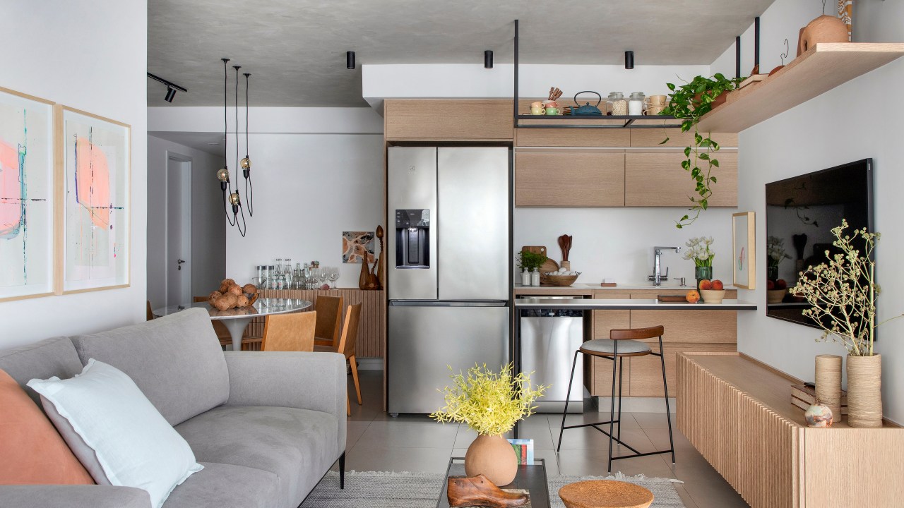Apê de 105 m² para mãe e filha tem dois espaços de estar e cozinha gourmet. Projeto de Rafael Ramos. Na foto, sala de estar, sofá cinza, cozinha integrada com bancada, prateleira suspensa, geladeira.