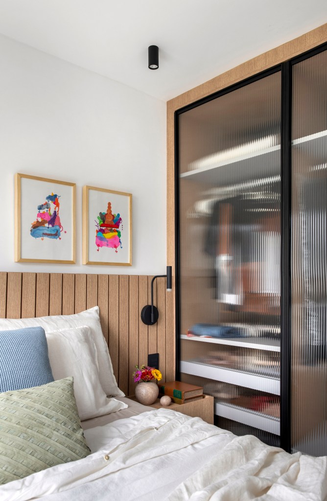 Apê de 105 m² para mãe e filha tem dois espaços de estar e cozinha gourmet. Projeto de Rafael Ramos. Na foto, quarto com cabeceira de marcenaria em lambris, quadros acima da cabeceira, armário com portas de vidro canelado.