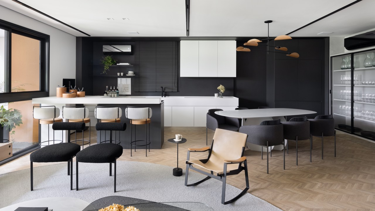 Apê de 142 m² tem paleta elegante de preto e branco e layout integrado. Projeto de Boher Arquitetos. Na foto, sala de estar integrada com cozinha, piso de madeira, tapete cinza, mesa de centro, cozinha com marcenaria branca, parede preta.