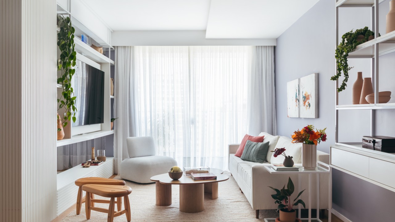 Apê de 160 m² em tons pastel alegres tem painel giratório de TV. Projeto de Ikeda Arquitetura. Na foto, sala de estar com parede lilás, painel giratório de tv, sofá branco, cortina, poltrona baixa branca.