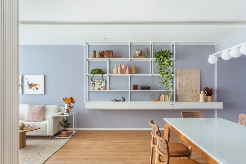 Apê de 160 m² em tons pastel alegres tem painel giratório de TV. Projeto de Ikeda Arquitetura. Na foto, sala de jantar com parede lilás, estante vazada branca com vasos e plantas.