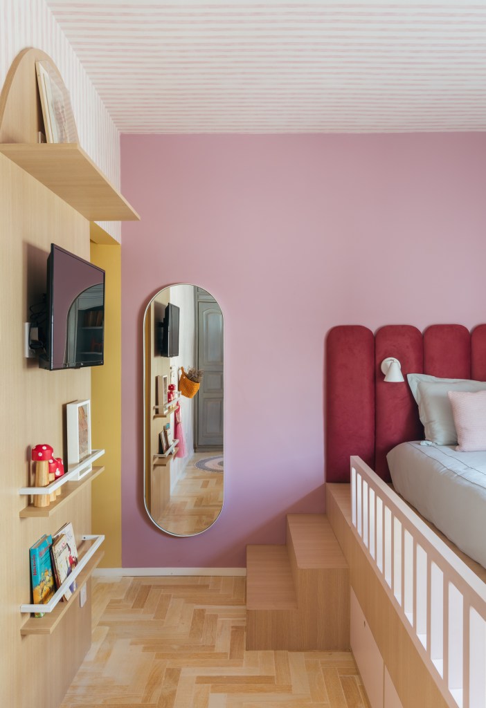 Apê de 160 m² em tons pastel alegres tem painel giratório de TV. Projeto de Ikeda Arquitetura. Na foto, quarto infantil com parede rosa, cabeceira estofada, cama em plataforma com escada, espelho de parede, painel com tv e prateleira para livros.