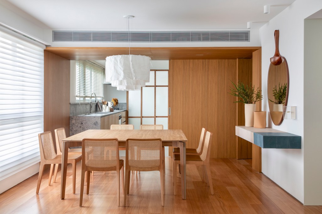 Apê de 180 m² com sala íntima equilibra minimalismo e conforto. Projeto de Studio LAK. Na foto, sala de jantar com piso, mesa e cadeiras em madeira, luminária de tecido, porta de correr de madeira integrando a cozinha.