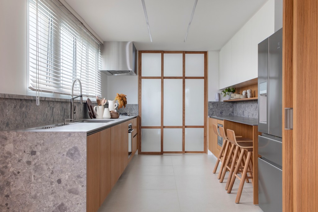Apê de 180 m² com sala íntima equilibra minimalismo e conforto. Projeto de Studio LAK. Na foto, cozinha com bancada em granilite, armários em tom madeira, bancada para refeições rápidas.