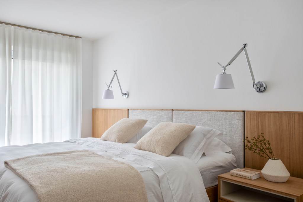 Apê de 180 m² com sala íntima equilibra minimalismo e conforto. Projeto de Studio LAK. Na foto, quarto de casal com cabeceira acolchoada em painel de marcenaria, luminárias de parede brancas.