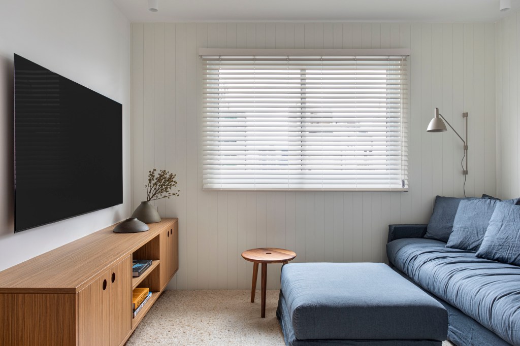 Apê de 180 m² com sala íntima equilibra minimalismo e conforto. Projeto de Studio LAK. Na foto, sala íntima com sofá azul, tv, persiana.
