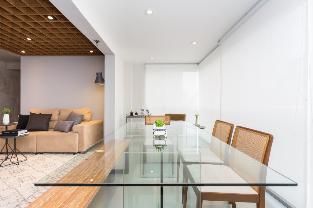 Apê de 89 m² ganha paredes com cimento queimado e piso com camada de epóxi. Projeto de Ju Matos. Na foto, varanda integrada com persianas brancas, mesa de vidro.
