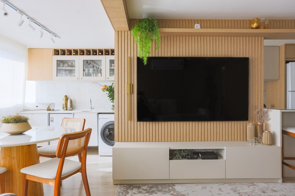 Bege, móveis planejados e muita madeira compõem o décor de apê de 95 m². Projeto de Thais Aguiar. Na foto, sala com varanda e lavanderia. Painel ripado de TV.