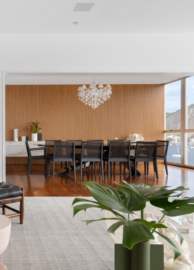 Caixa de marcenaria envolve área de estar e mimetiza portas em apê. Projeto de Studio Lak. Na foto, sala de jantar com parede com painel ripado, mesa e cadeiras pretas, lustre.