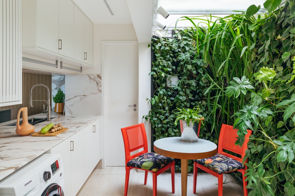 Cobertura tem sauna com paredes de vidro e área gourmet com balanços. Projeto de Vivian Reimers. Na foto, área de refeições, mesa redonda pequena, cadeiras laranja, jardim vertical.