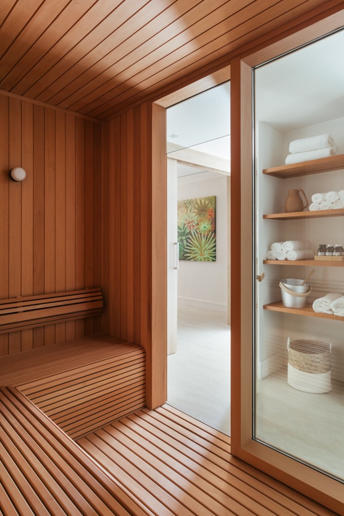 Cobertura tem sauna com paredes de vidro e área gourmet com balanços. Projeto de Vivian Reimers. Na foto, sauna revestida de madeira com banco, porta e parede de vidro.