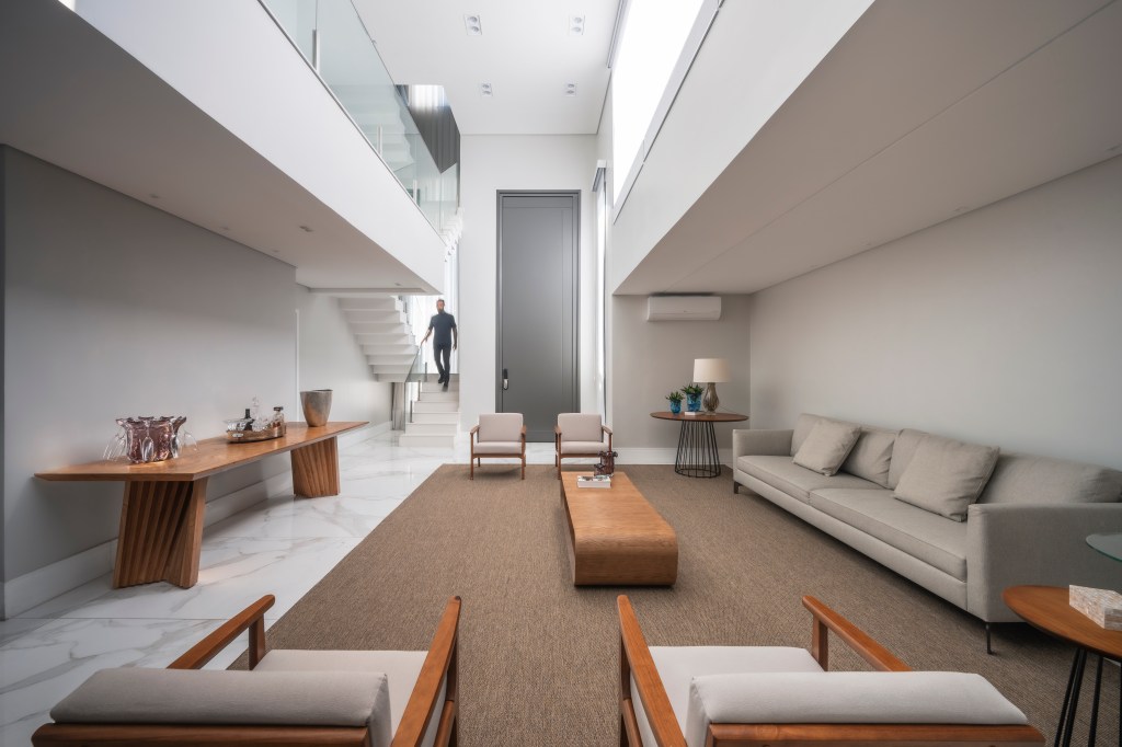 Com 500 m², casa clean tem paleta clara, closet e cozinha provençal. Projeto de PB Arquitetura. Na foto, sala de estar com pé-direito alto, tapete bege, sofá bege, poltronas brancas, mezanino.
