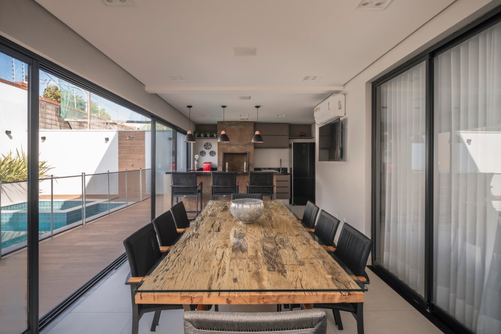 Com 500 m², casa clean tem paleta clara, closet e cozinha provençal. Projeto de PB Arquitetura. Na foto, área gourmet com churrasqueira, mesa de madeira rústica, cadeiras pretas, bancada com banquetas.