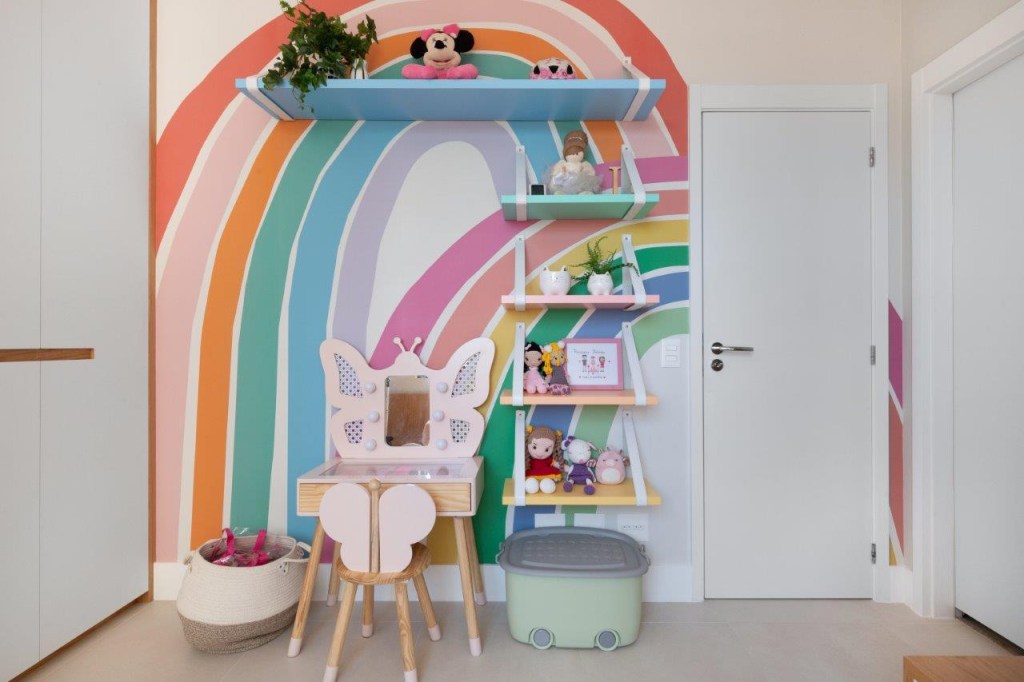 Cores verde, azul, rosa e detalhes em pedra e palha pontuam apê de 180 m². Projeto de Natália Salla. Na foto, quarto infantil com arco-íris pintado na parede, penteadeira e cadeira de borboleta.