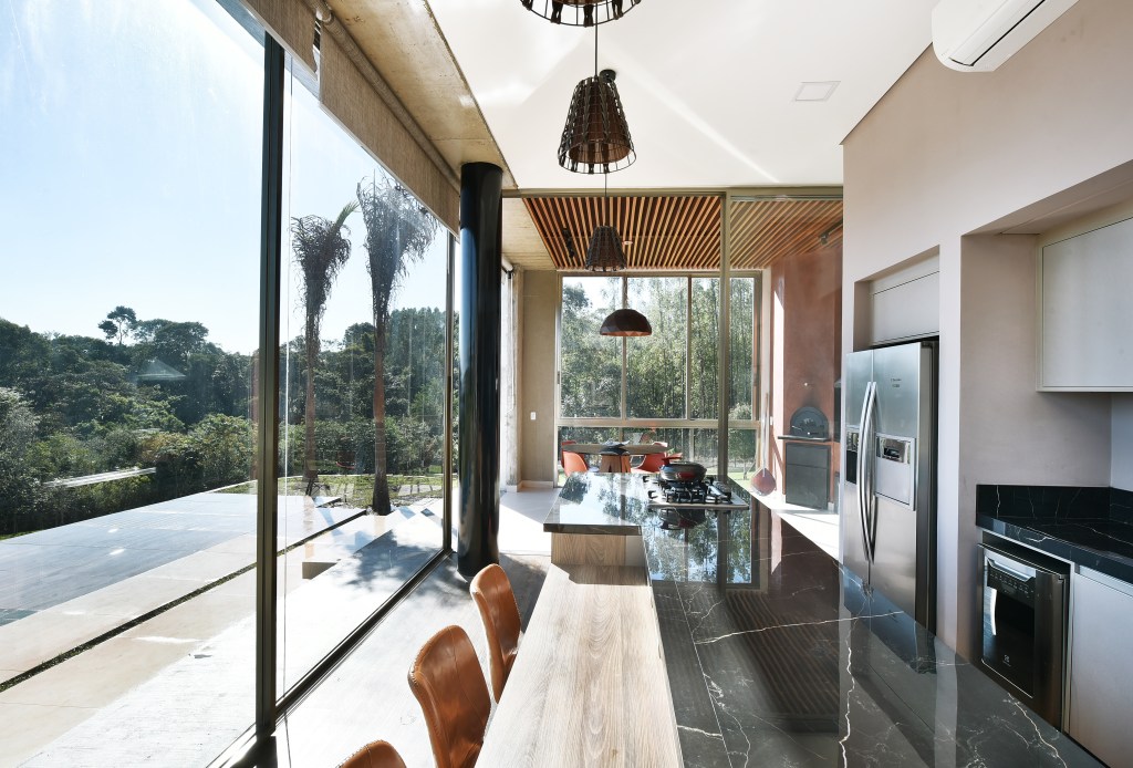 Grandes aberturas realçam o décor clean desta casa de veraneio com piscina. Projeto de Marcela Rocca. Na foto, cozinha com grandes aberturas de vidro, ilha com bancada de granito preto.