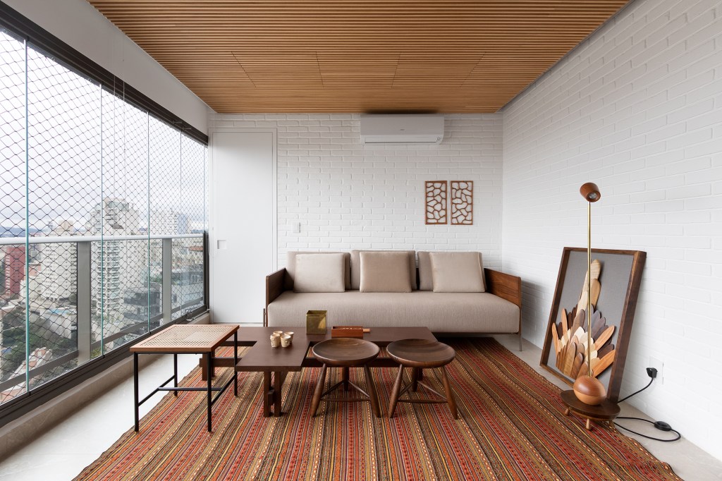 Linguagem neutra e atemporal definem o décor de apartamento de 140 m². Projeto MNBR Arquitetos. Na foto, varanda com parede de tijolo, tapete e quadro.