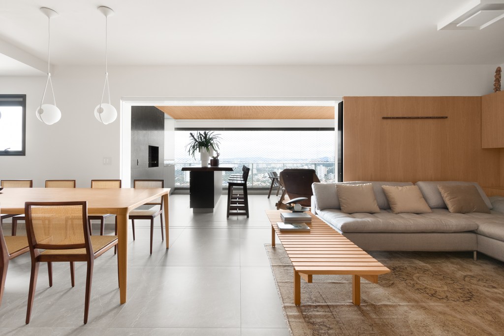 Linguagem neutra e atemporal definem o décor de apartamento de 140 m². Projeto MNBR Arquitetos. Na foto, sala integrada com a varanda e jantar. Paredes de madeira e tapete.