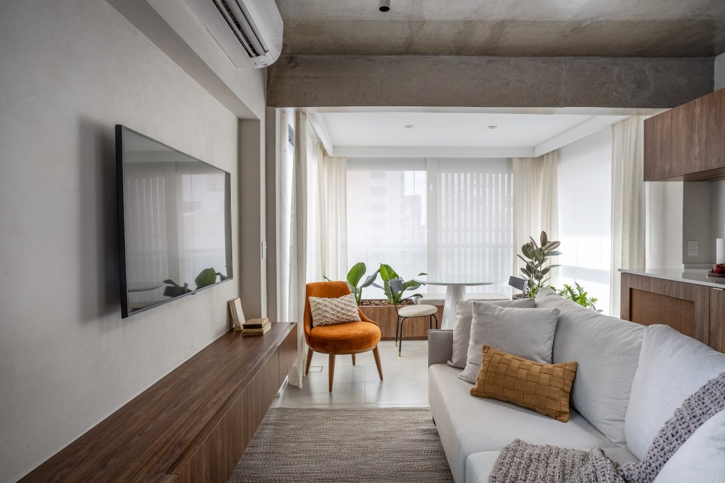 Marcenaria personalizada otimiza espaço e esconde eletros em apê de 48 m². Projeto de Carolina Gava. Na foto, sala de estar, laje de concreto aparente, poltrona laranja.