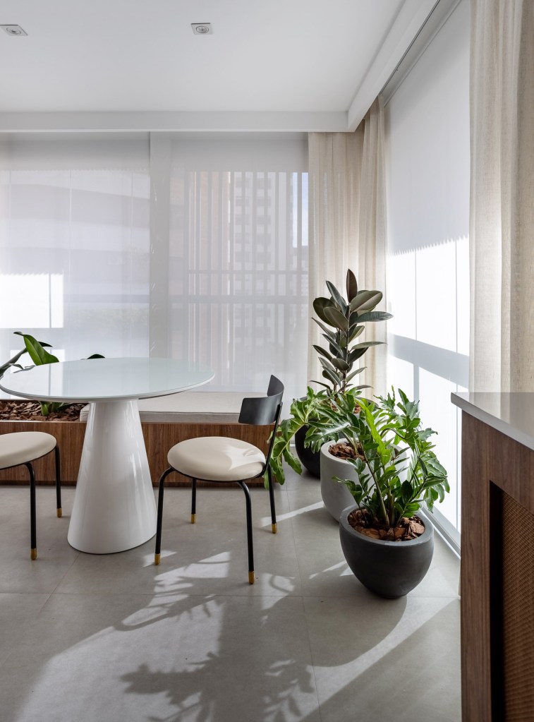 Marcenaria personalizada otimiza espaço e esconde eletros em apê de 48 m². Projeto de Carolina Gava. Na foto, sala de jantar, mesa redonda branca pequena, plantas, piso de porcelanato.