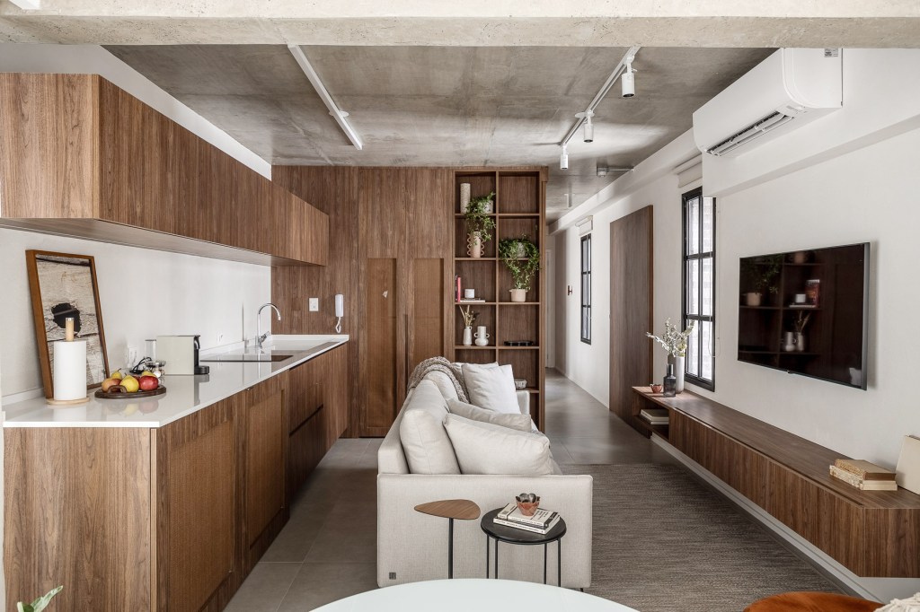 Marcenaria personalizada otimiza espaço e esconde eletros em apê de 48 m². Projeto de Carolina Gava. Na foto, sala integrada com cozinha, sofá branco, piso de porcelanato.