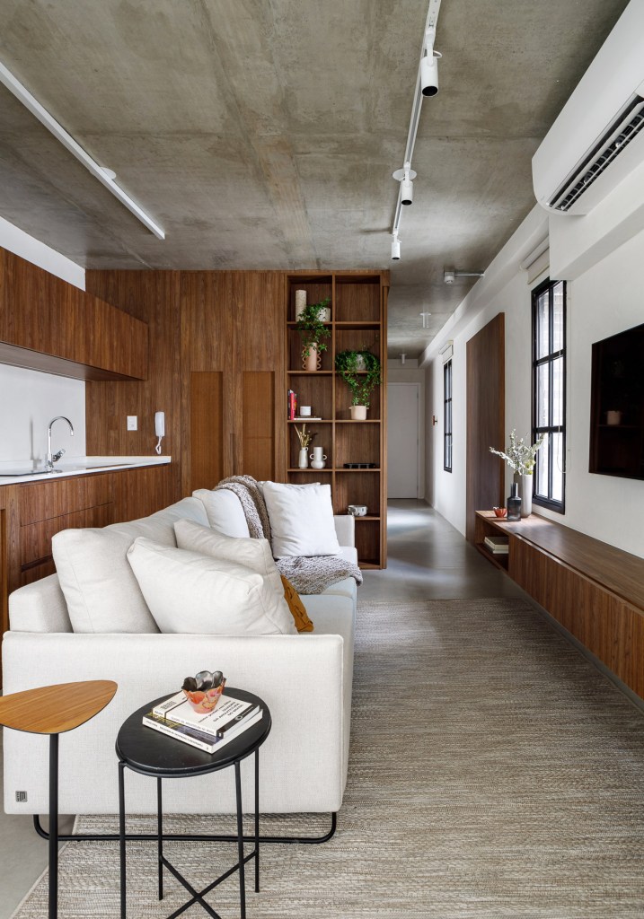 Marcenaria personalizada otimiza espaço e esconde eletros em apê de 48 m². Projeto de Carolina Gava. Na foto, sala integrada com cozinha, sofá branco, madeira escura, piso de porcelanato.