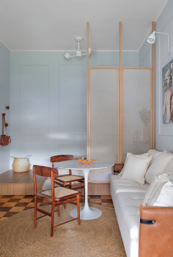 Descubra microapê rococó de apenas 30 m² no hotel-cassino Quitandinha. Projeto de Manuela Fleck. Na foto, sala de estar pequena, sofá branco, divisória transparente, mesa redonda pequena, cadeiras de madeira, piso de madeira.