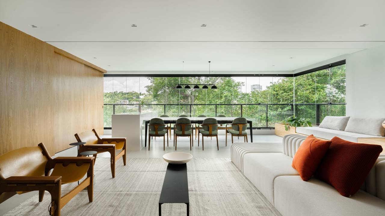 Painel de marcenaria abriga adega, TV e oculta portas neste apê de 170 m². Projeto Estudio Pluri. Na foto, sala de estar e varanda integradas com sofá e mesa.