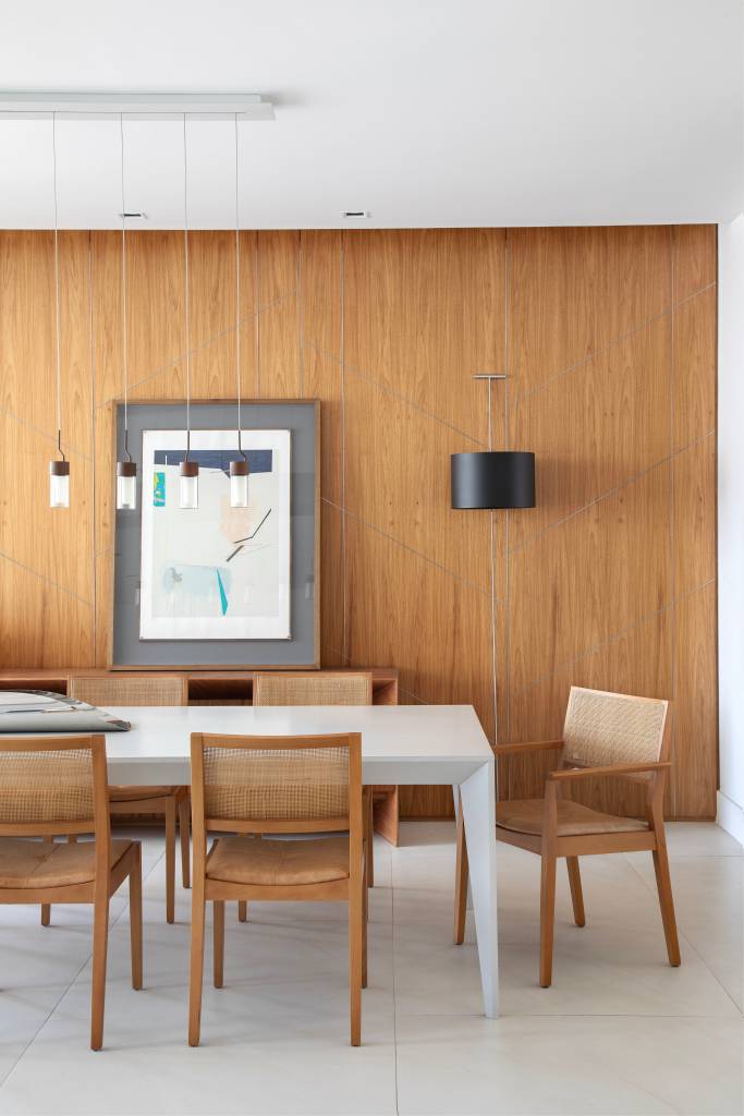 Quarto deste apê tem grande painel de freijó natural com lâminas diagonais. Projeto de Cristina Bezamat. Na foto, sala de jantar com parede revestida com painel de madeira e mesa grande branca.
