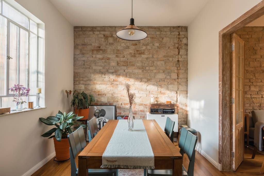 Sobrado de 100 m² ganha solarium e décor que é mix de rústico e retrô. Projeto de Palladino Arquitetura. Na foto, sala de jantar com mesa de madeira, parede de tijolinhos e plantas.