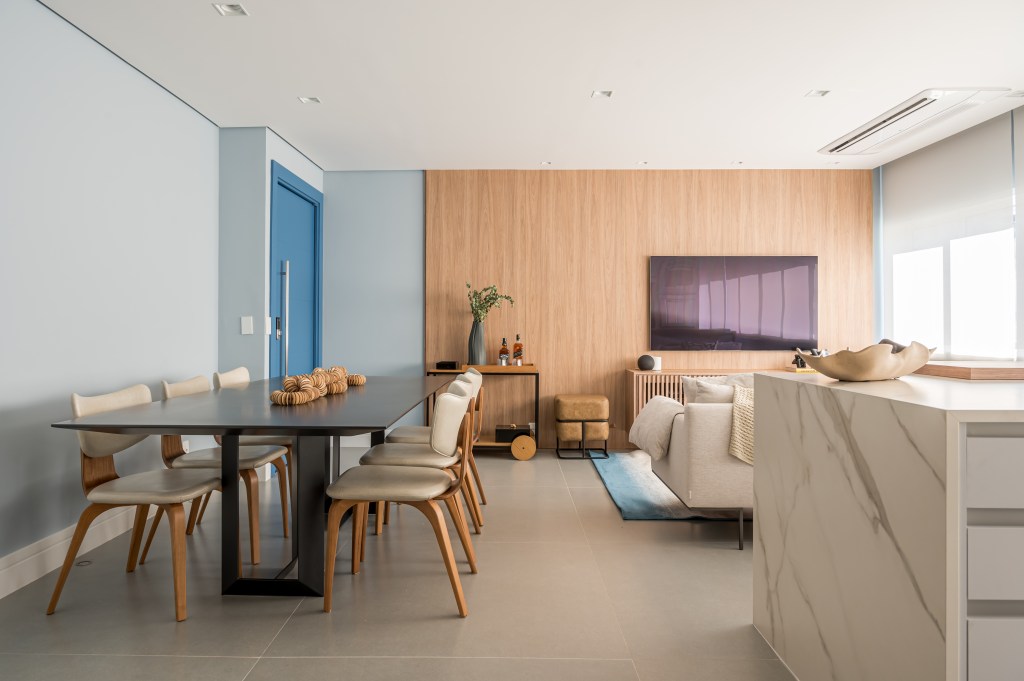 Tons de azul e integração total na área social marcam apê de 107 m². Projeto de Daniela Funari. Na foto, sala integrada, piso de porcelanato, mesa preta, cadeiras, painel de madeira.