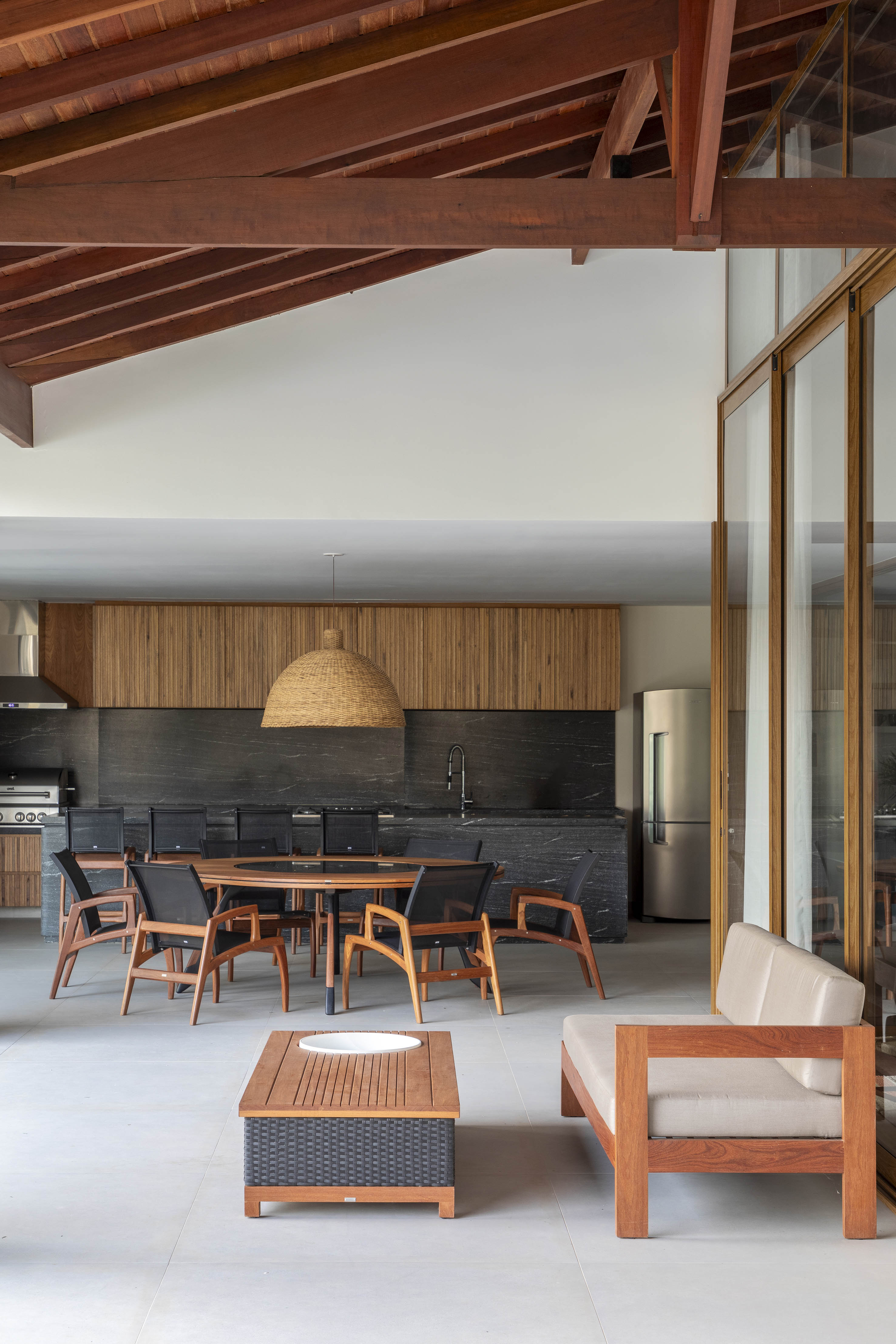 Tons naturais e base neutra marcam casa de campo com cozinha provençal. Projeto de João Panaggio. Na foto, varanda gourmet com mesa de madeira redonda, bancada preta e marcenaria preta.