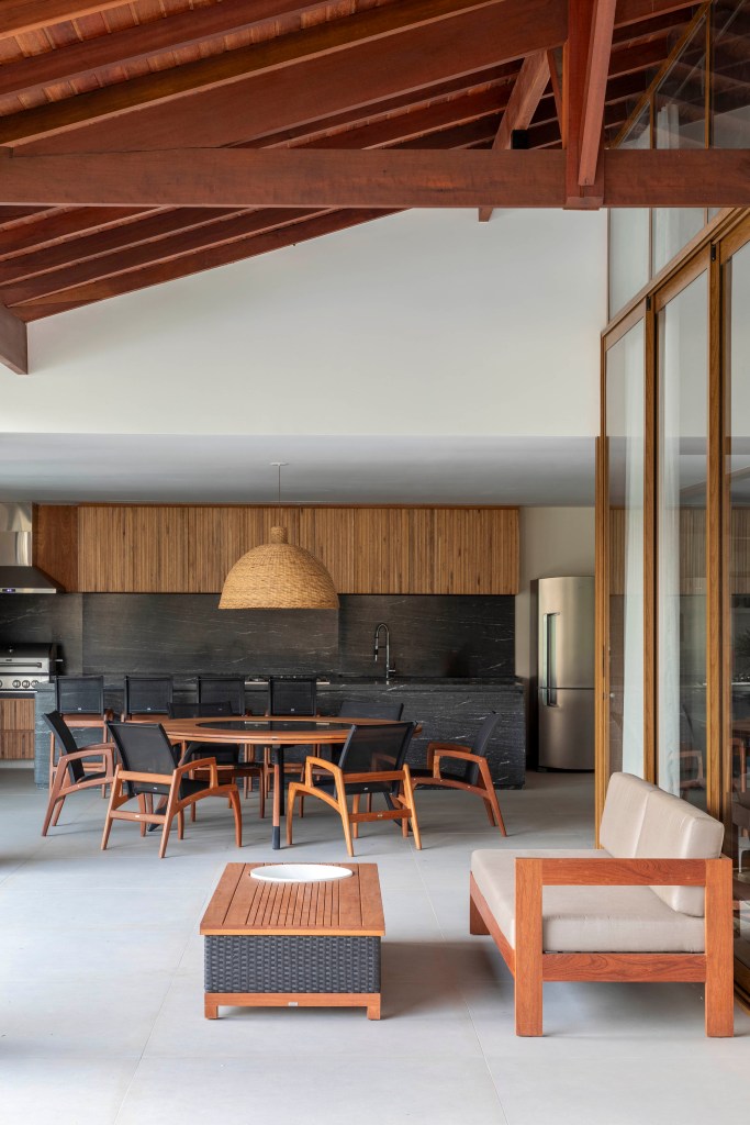 Tons naturais e base neutra marcam casa de campo com cozinha provençal. Projeto de João Panaggio. Na foto, varanda gourmet com mesa de madeira redonda, bancada preta e marcenaria preta.