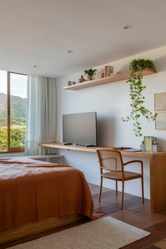 Tons naturais e base neutra marcam casa de campo com cozinha provençal. Projeto de João Panaggio. Na foto, quarto de casal com aparador de madeira com tv e home office. Prateleira com plantas.