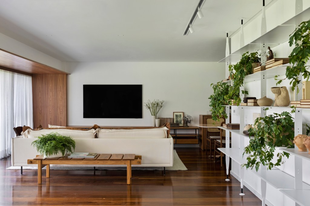 Apê integrado ganha décor contemporâneo com muita marcenaria em freijó. Projeto de João Panaggio. Na foto, sala de estar, tv, piso de madeira, prateleira de plantas, sofá off white.