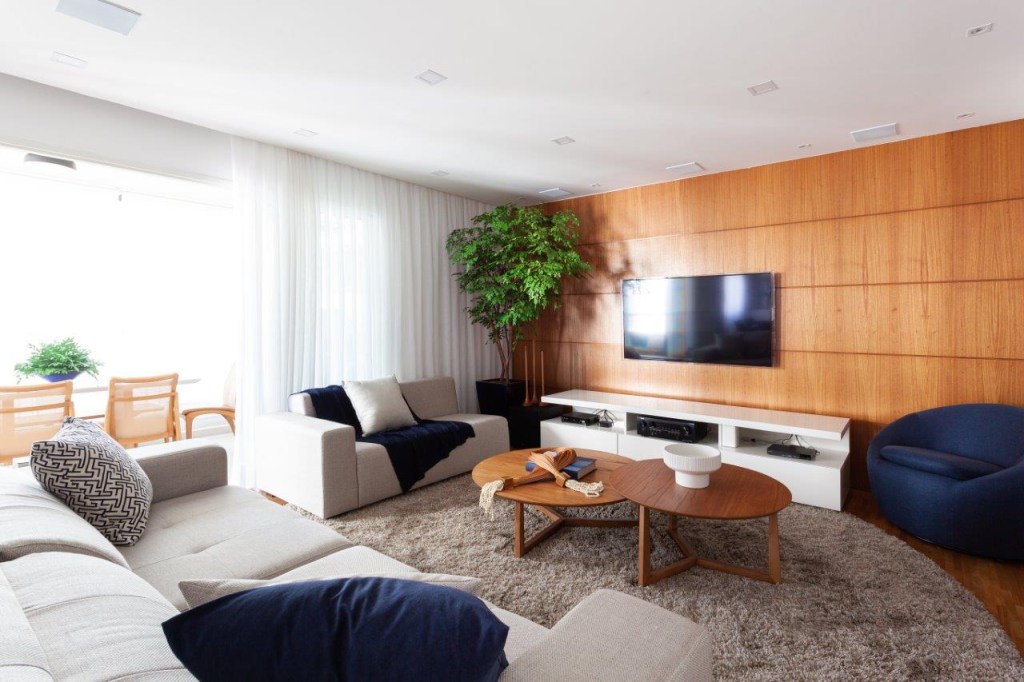 Base neutra com pontos marcantes em azul definem apê de 167 m². Projeto de Ju Miranda. Na foto, sala de estar, tv, parede com painel de madeira, mesa de centro de madeira.