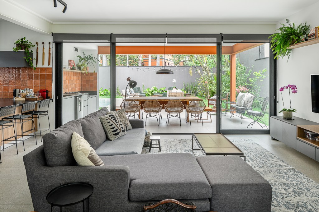 Casa de 415 m² com mezanino e varanda gourmet é inspirada em chalé alemão. Projeto de Arkitito Arquitetura. Na foto, sala de estar, portas de correr de vidro para varanda, sofá cinza.