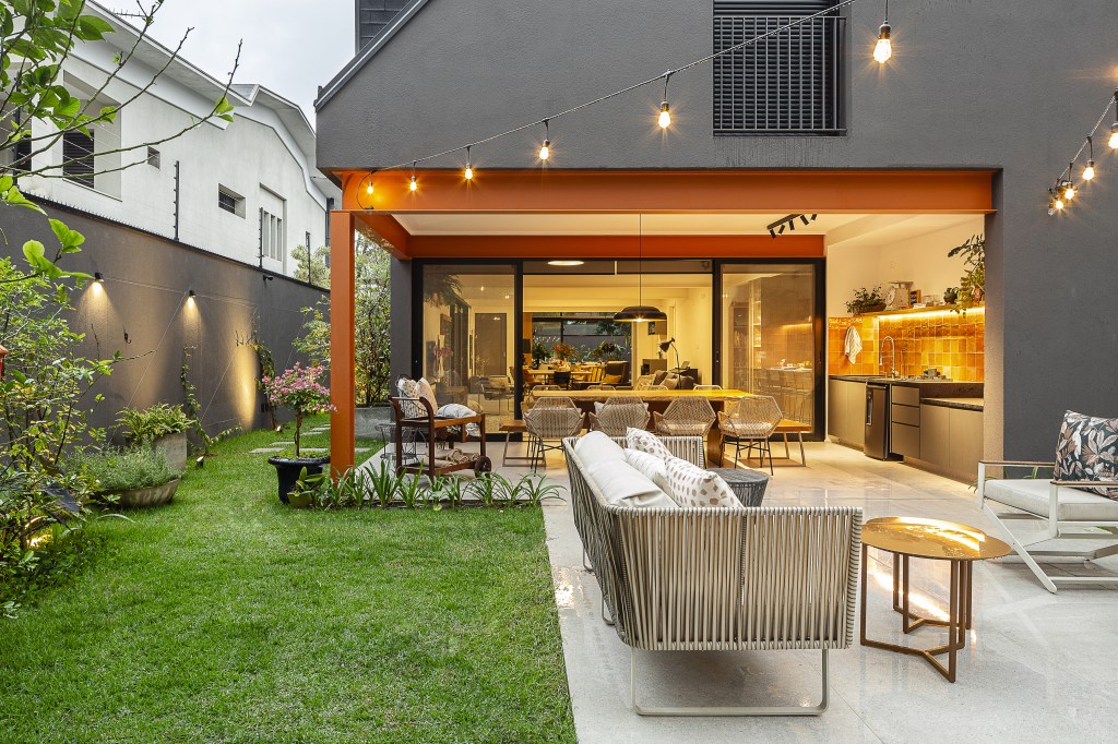 Casa de 415 m² com mezanino e varanda gourmet é inspirada em chalé alemão. Projeto de Arkitito Arquitetura. Na foto, área externa com jardim, pergolado iluminado, sofás, varanda gourmet.
