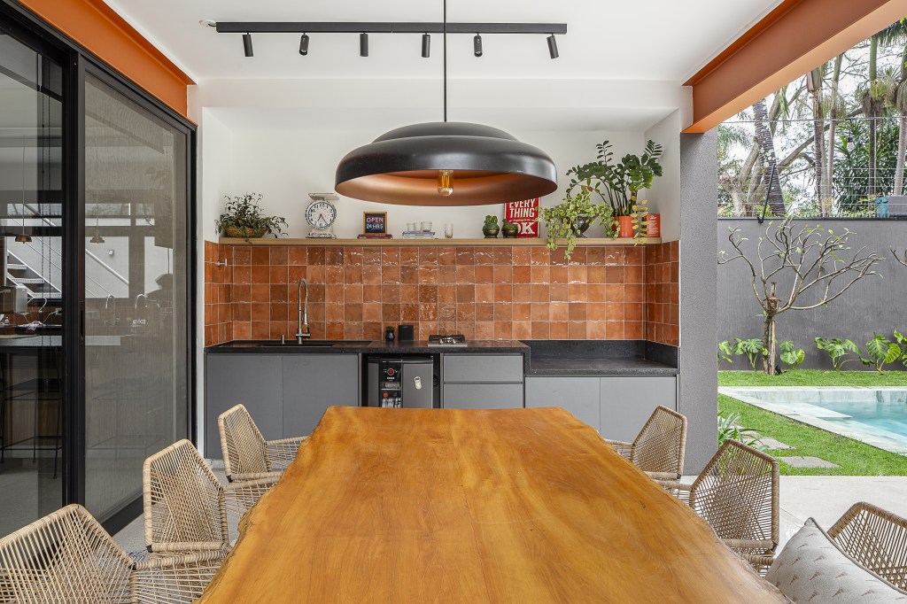 Casa de 415 m² com mezanino e varanda gourmet é inspirada em chalé alemão. Projeto de Arkitito Arquitetura. Na foto, varanda gourmet, parede com azulejos terracota, mesa de madeira, bancada preta.