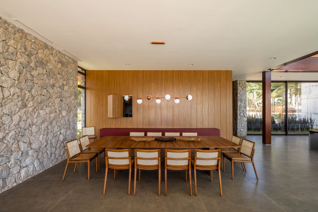 Casa é composta por três volumes ligados por pergolado de madeira. Projeto de Arkitito. Na foto, sala de jantar, mesa de madeira, parede de pedra.