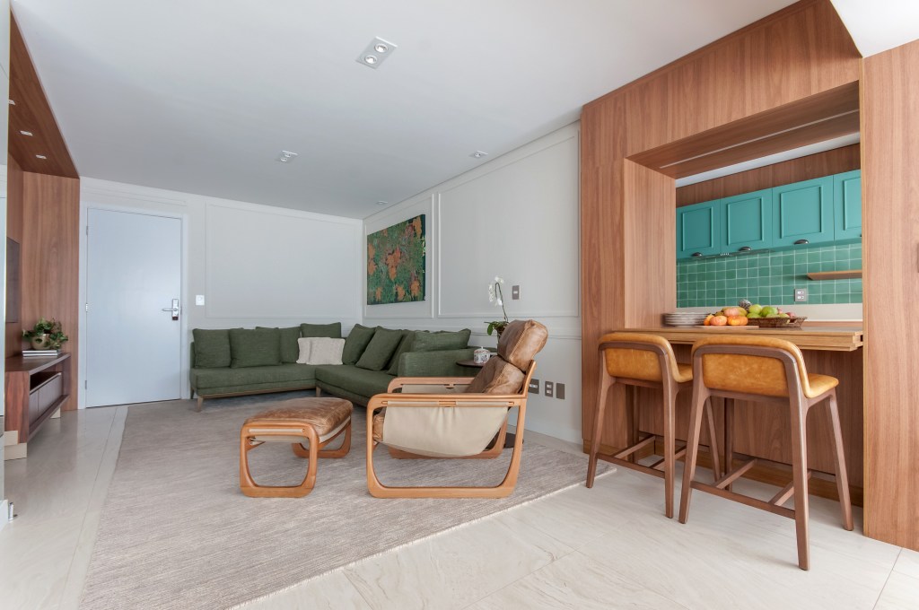 Cozinha com armários e revestimentos verde azulados é o charme deste apê. Projeto de Paiva e Passarini. Na foto, sala de estar com sofá verde L, poltrona com pufe, pórtico de madeira com abertura para cozinha.