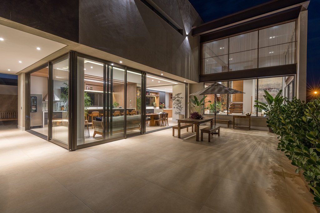 Grandes vãos de vidro integram esta casa de 600 m² à natureza do entorno. Projeto Beth Araújo. Na foto, varanda iluminada com ombrelone.