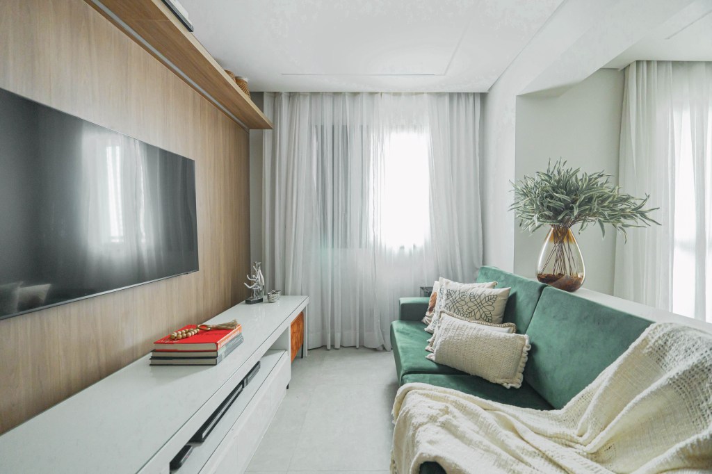 Layout e revestimentos de apê de 90 m² foram pensados para casal de idosos. Projeto de Larissa Albuquerque. Na foto, sala de estar, parede revestida de madeira, sofá verde, rack branco.