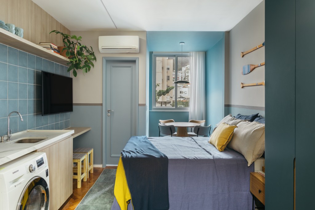 Microapê com 25 m² ganha tons azuis com reforma de apenas 3 meses. Projeto de Rodolfo Consoli. Na foto, loft com paredes azuis, lavanderia, cozinha, bancada para tv, mesa de jantar pequena, cama de casal.