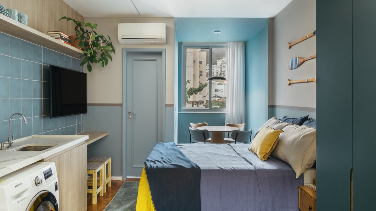 Microapê com 25 m² ganha tons azuis com reforma de apenas 3 meses. Projeto de Rodolfo Consoli. Na foto, loft com paredes azuis, lavanderia, cozinha, bancada para tv, mesa de jantar pequena, cama de casal.