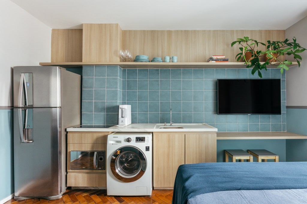 Microapê com 25 m² ganha tons azuis com reforma de apenas 3 meses. Projeto de Rodolfo Consoli. Na foto, loft com parede de revestimento cerâmico azul abrigando lavanderia, cozinha e tv. Máquina de lavar.