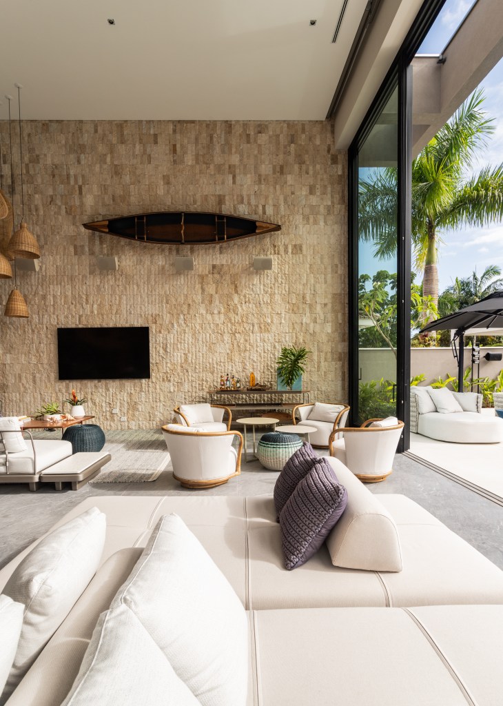Mix de elementos artesanais, naturais e tecnologia definem casa de praia. Projeto de Patricia Penna. Na foto, sala com pé direito duplo, parede de pedra, grandes janelas, sofás claros.