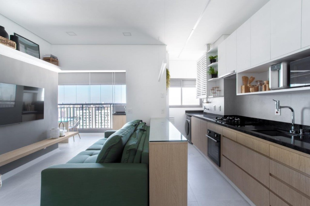 Sofá retrátil verde é destaque na sala de estar deste apartamento de 67 m². Projeto de Ju Miranda. Na foto, cozinha integrada, marcenaria branca, bancada preta, sofá verde na sala.