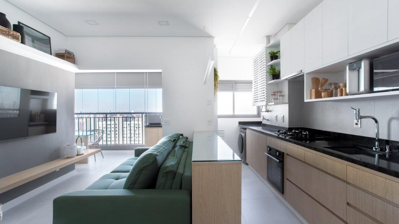 Sofá retrátil verde é destaque na sala de estar deste apartamento de 67 m². Projeto de Ju Miranda. Na foto, cozinha integrada, marcenaria branca, bancada preta, sofá verde na sala.