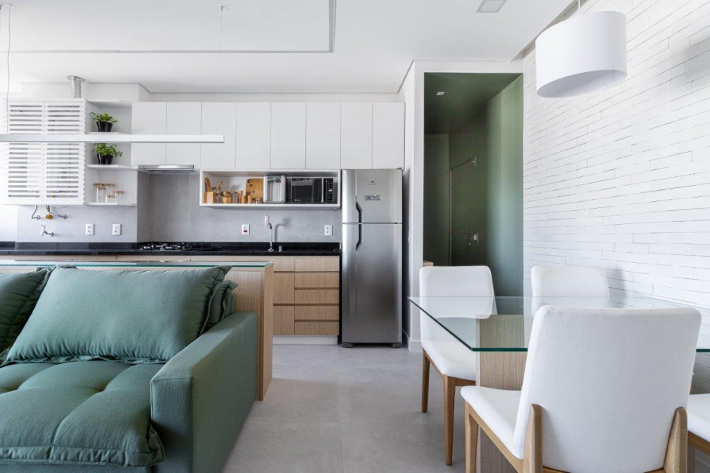Sofá retrátil verde é destaque na sala de estar deste apartamento de 67 m². Projeto de Ju Miranda. Na foto, sala de estar integrada com cozinha, sofá verde, hall de entrada verde, sala de jantar com mesa com tampo de vidro.